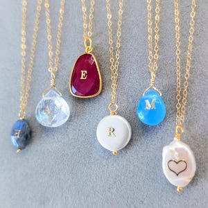 Puede incluir: Una colección de collares dorados con varios colgantes de piedras preciosas. Los colgantes incluyen un óvalo azul oscuro, un cristal transparente, una piedra roja con la letra "E", una perla blanca con la letra "R", una lágrima azul con la letra "M" y una perla blanca con un corazón.