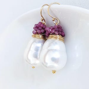 May include: A pair of elegant earrings featuring large, pearlescent white baroque pearls. The pearls are accented with clusters of small, deep pink gemstones and wrapped with gold wire. Gold-colored ear wires complete the design.