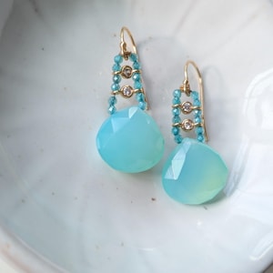 May include: A pair of gold drop earrings featuring faceted light blue teardrop stones suspended from gold wires. Each earring includes two small clear round stones flanked by rows of tiny blue beads, finished with a gold hook ear wire.