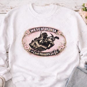 May include: White sweatshirt featuring a pink oval design with the words "West Virginia" and "Coal Miners Wife." The design includes a silhouette of a coal miner and floral accents. The sweatshirt is paired with jeans and sneakers.