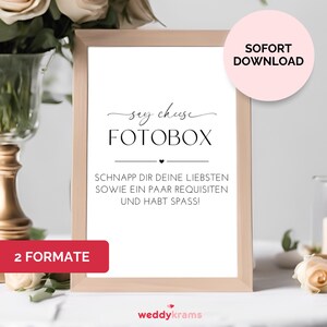 Puede incluir: Un cartel blanco con texto negro que dice "say cheese FOTOBOX" y "SCHNAPP DIR DEINE LIEBSTEN SOWIE EIN PAAR REQUISITEN UND HABT SPASS!" El cartel está en un marco de madera y está sentado en una mesa blanca con flores y una vela.