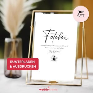 Op de afbeelding: Een gouden lijst met de tekst "Fotobox" in zwarte cursieve letters. Het bord bevat ook instructies voor het gebruik van een fotohokje, met de tekst "Schnapp dir ein paar Requisiten, stell dich vor die Kamera und drucke den Auslöser. Say Cheese!" in zwarte tekst. Het bord staat op een wit oppervlak met een witte vaas en een gedroogde plant op de achtergrond.