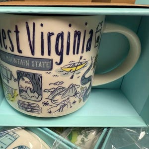 May include: White ceramic mug with a handle, featuring the words "West Virginia" in navy blue, along with illustrations of the state's landmarks. The mug is packaged in a light blue box, perfect for gifting.