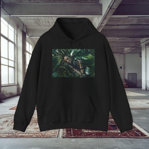 May include: Black hoodie with a rectangular graphic. The graphic shows a person amongst green leaves, wearing silver jewellery. The person has their eyes closed. The background is a concrete industrial space with large windows and rugs.