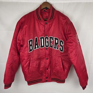 May include: A bright red satin varsity jacket featuring the word BADGERS in bold black and white lettering across the chest. The jacket includes a snap-button front closure, a ribbed collar with black stripes, and matching ribbed cuffs and hem.