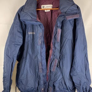 May include: A navy blue Columbia Sportswear jacket with a zipper closure and a drawstring at the bottom. The jacket has a burgundy lining and a white Columbia logo on the left chest.
