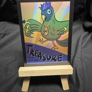 May include: A colorful illustration of a parrot wearing a crown and jewelry, with the word "TREASURE" at the bottom. The artwork is displayed on a small wooden easel, set against a dark background.