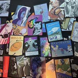 May include: A collection of rectangular game cards featuring diverse illustrations including a goose labeled Bird, a coin labeled Trigger, a character labeled Copy, and various fantasy figures. The cards display vibrant colors and text.