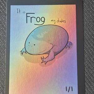 Op de afbeelding: Een rechthoekige verzamelkaart met een zwarte rand en een holografische regenboogachtergrond. De kaart bevat een tekening van een ronde, groene kikker en de tekst "It is Frog my dudes" bovenaan, met "1/1" rechtsonder gedrukt.