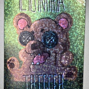 May include: A card with a glittery green background featuring a cartoon bear with button eyes and a button nose. The bear has a pink tongue sticking out. The words "ILUMIRA" and "TRIGGER" are written on the card.