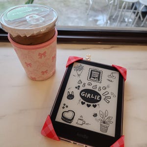 May include: A Kindle e-reader with a decorative cover featuring doodles and the word "GIRLIE". The e-reader is protected by red and pink gingham corner protectors. A pink cup sleeve with bow patterns holds a coffee drink.