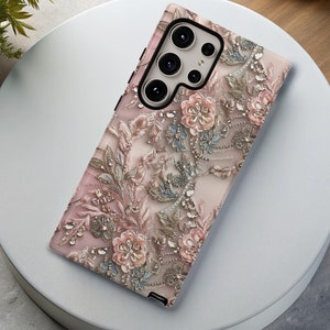 Puede incluir: Una funda para teléfono rosa y dorada con flores y un recorte para la cámara. La funda presenta un delicado diseño floral con pedrería y un fondo texturizado.