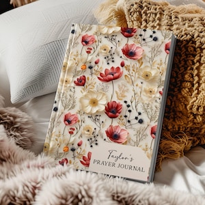 May include: A floral-patterned journal with red and cream-colored flowers and the text "Taylor's Prayer Journal." The journal is lying on a bed with pillows and a blanket. The cover features a variety of flowers and foliage.