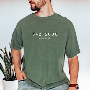 Puede incluir: Una camiseta verde con el texto "5+2=5000" y "JOHN 6:9-13" impreso en la parte delantera.