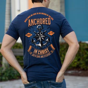 Puede incluir: Camiseta azul marino con un diseño gráfico en la espalda. El diseño presenta un ancla, una rosa de los vientos y el texto "ANCHORED IN CHRIST...BOTH SURE AND STEDFAST." El texto adicional incluye "WHICH HOPE WE HAVE AS AN ANCHOR OF THE SOUL" y "HEB 6:19."