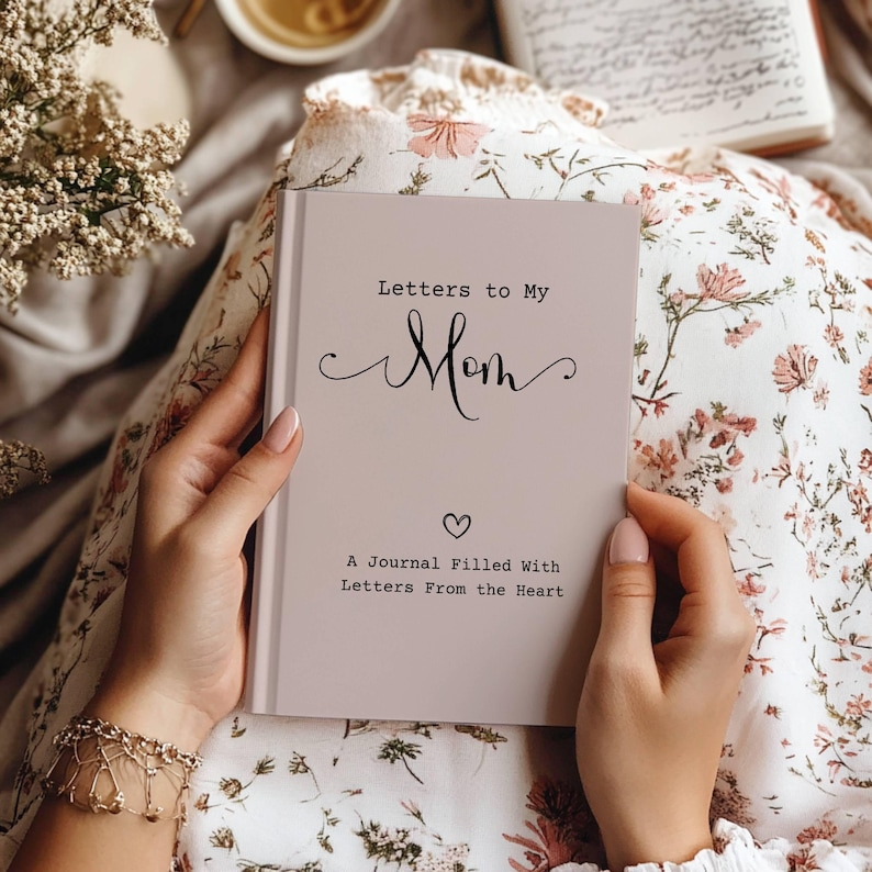 May include: A light pink journal titled "Letters to My Mom" with a heart symbol. The journal is held open, revealing the cover text. The background includes a floral patterned fabric and a cup of tea.