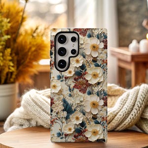 May include: A white phone case with a floral design. The flowers are white and have a 3D effect. The background is a light beige color.