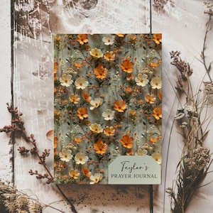 May include: A floral prayer journal with a light blue background and a pattern of orange and white flowers. The journal has the text "Taylor's PRAYER JOURNAL" on the front cover.