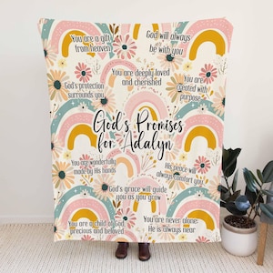 以下が含まれることがあります： 虹と花の模様が描かれた白と黄色の毛布。毛布には「God's Promises for Adalyn」という文字と、いくつかの励みになる言葉が書かれています。