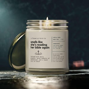 Puede incluir: Una vela blanca en un tarro de vidrio con tapa dorada. La vela está etiquetada con "A Thread of Faith Co" y "smells like she's reading her bible again". La vela está infundida con oraciones diarias, momentos de reflexión tranquilos y un corazón lleno de fe. La vela tiene aroma a vainilla y tiene un tiempo de combustión de 42 horas.
