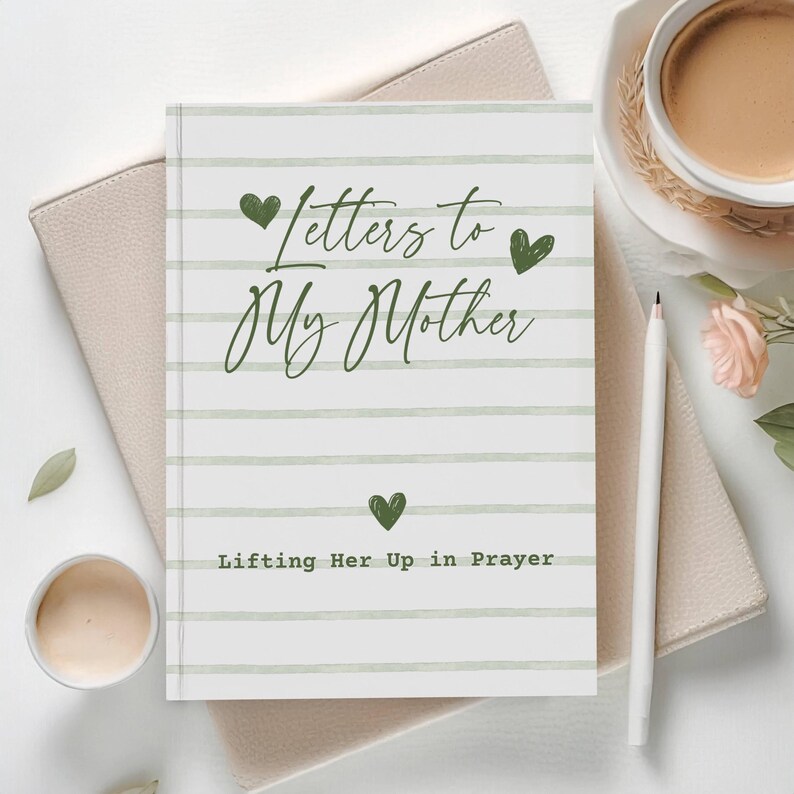 May include: A journal titled "Letters to My Mother" with green heart accents and text "Lifting Her Up in Prayer." The notebook is open on a light-colored surface, next to a cup of coffee, a pen, and a small flower arrangement.