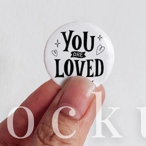 May include: A circular white pin button with black text that reads YOU ARE LOVED, accented by small heart and star icons. The button has a diameter of 1.25 inches as indicated by the text above it. The design is simple, clean, and high-contrast.