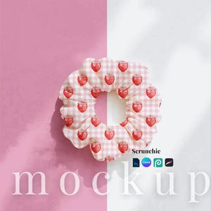 May include: A scrunchie with a pink and white gingham pattern, decorated with red strawberry motifs. The accessory is set against a two-tone backdrop of pink and white. The word "mockup" is visible at the bottom.