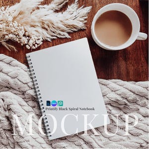 May include: A white spiral notebook mockup on a white knitted blanket with a cup of coffee and dried flowers. The notebook has the text "Printify Black Spiral Notebook" printed on the cover.