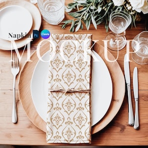 May include: A white dinner plate with a gold patterned napkin on top of it. The plate is on a wooden table with a wooden charger plate underneath. There are two silver forks and a silver knife on the table.