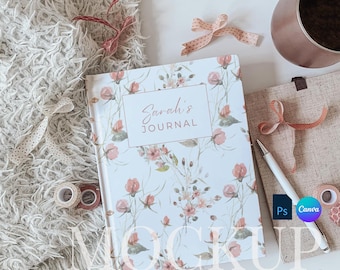 May include: A hardcover journal with a floral design and the text "Sarah's JOURNAL" on the front cover. The journal is on a white surface with a fluffy blanket, ribbon, and a cup of coffee.