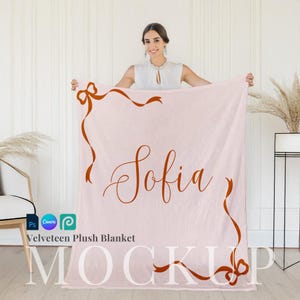 May include: A pink velvety plush blanket with a brown ribbon design and the name "Sofia" written in cursive. The blanket is being held by a person in a white shirt.
