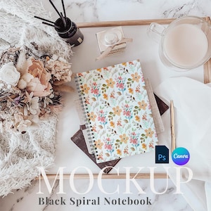 Puede incluir: Un cuaderno espiral blanco con un patrón floral en la portada. El cuaderno está en una bandeja de madera con otros artículos decorativos, incluyendo un vaso de leche, una pequeña botella de aceite esencial y una caja decorativa. El texto "MOCKUP" y "Black Spiral Notebook" está impreso en la bandeja.