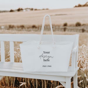 May include: White tote bag with the text "Your design here" and "PSD | PNG" on a white bench in a field of tall grass.