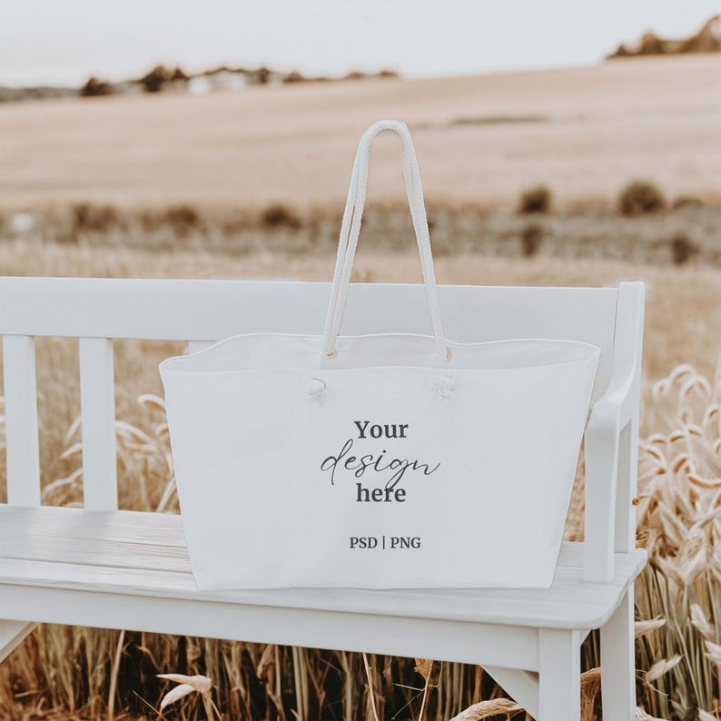 May include: White tote bag with the text "Your design here" and "PSD | PNG" on a white bench in a field of tall grass.