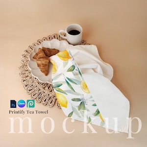 Pode incluir: Toalha de chá branca com um design de limão e folhas. A toalha está dobrada e colocada em um tapete de vime com um prato branco e dois croissants. Uma xícara de café branca está no tapete de vime. O texto "Printify Tea Towel" está abaixo da imagem.