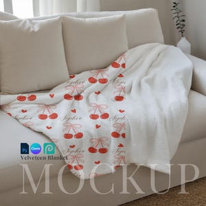 May include: A white velvety blanket with a red cherry pattern and the name "Sophia" repeated throughout. The blanket is folded and draped over a white couch with pillows.