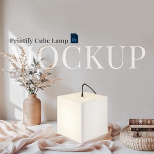 Op de afbeelding: Mockup van een witte kubuslamp op een tafel met een vaas met gedroogde bloemen en een stapel boeken. De lamp is aan en geeft een warm licht. De tekst "Printify Cube Lamp Ps" is zichtbaar in de linkerbovenhoek van de afbeelding.