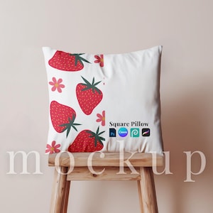 May include: White square pillow with a pattern of red strawberries and pink flowers. The pillow is displayed on a wooden stool. Text on the pillow reads "Square Pillow" with design software logos. The word "mockup" is at the bottom.