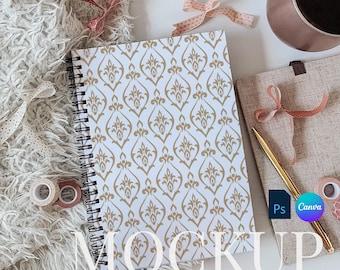 May include: A white spiral notebook with a gold damask pattern. The notebook is on a white surface with a fuzzy white blanket, a cup of coffee, and a brown burlap bag.