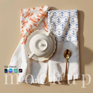 May include: Two white tea towels with nautical designs. One features orange lobsters, the other blue anchors. A white teapot and saucer with gold trim sit atop the towels, next to a gold spoon. The words "Tea Towel" are in the bottom left corner.