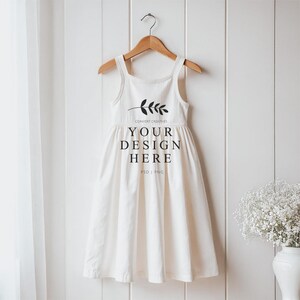 May include: White sleeveless dress with a gathered skirt hanging on a wooden hanger against a white wood panel background. The dress has a small sprig of leaves and the text "CONVERT CREATIVES YOUR DESIGN HERE PSD | PNG" printed on the front.