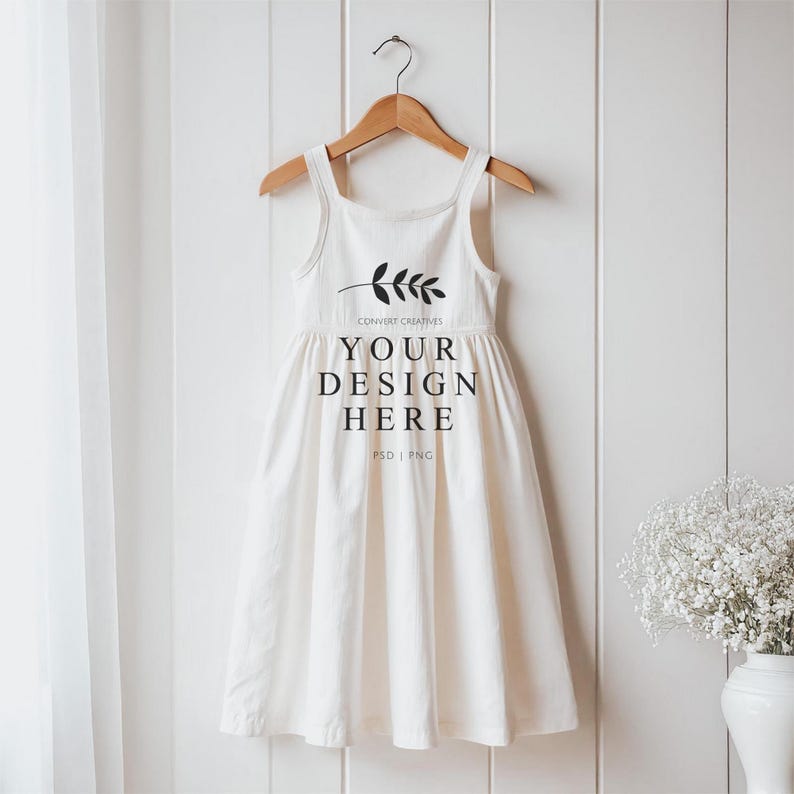 May include: White sleeveless dress with a gathered skirt hanging on a wooden hanger against a white wood panel background. The dress has a small sprig of leaves and the text "CONVERT CREATIVES YOUR DESIGN HERE PSD | PNG" printed on the front.