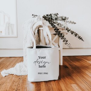 K&ouml;nnte beinhalten: Wei&szlig;e Canvas-Tragetasche mit schwarzen Griffen und dem Aufdruck "Your design here PSD | PNG" auf der Vorderseite.