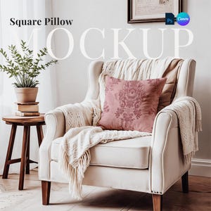 Op de afbeelding: Een witte fauteuil met een roze vierkant kussen met een bloemenpatroon. De stoel staat in een woonkamer met een bijzettafel en een plant.
