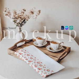 May include: A white tea towel with a red floral pattern is folded on a wooden tray with two cups of tea and saucers. The tray is on a white table with a vase of white flowers.