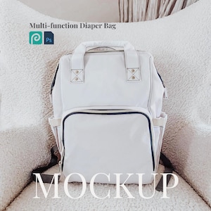 Op de afbeelding: Witte luiertas mockup met zwarte banden en ritsaccenten. De tas ligt op een wit, pluche oppervlak. De tekst "MOCKUP" is in wit op het oppervlak gedrukt.