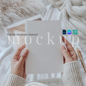 Könnte beinhalten: Ein weißes Hardcover-Journal mit dem Text "Printify Hardcover Journal" auf dem vorderen Umschlag. Das Journal wird von den Händen einer Person gehalten, im Hintergrund sind eine weiße Decke und ein flauschiger weißer Überwurf zu sehen.