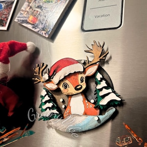 Puede incluir: Adorno navideño de madera con un ciervo que lleva un gorro de Papá Noel rojo, de pie en una escena de bosque nevado. El adorno está pintado en marrón, rojo, blanco y verde.