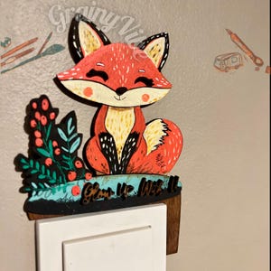 Op de afbeelding: Een houten lichtschakelaar met een geschilderd vos-ontwerp. De vos is rood, oranje en geel met zwarte accenten. De vos zit op een groene en blauwe achtergrond met rode bessen. De tekst "Glow Up, Love It" staat op de afdekplaat.
