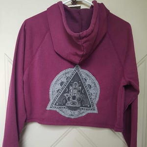 May include: A cropped, magenta hoodie with a drawstring hood. The back features a gray, circular design with a triangle at the center, containing an eye, hand, and other symbols. The hoodie has long sleeves and ribbed cuffs.